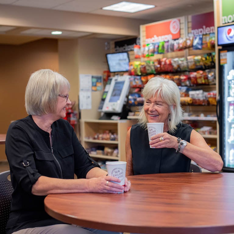 two women sitting at a table smiling at each other drinking new england coffee